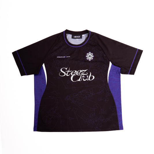 Un maillot noir et violet de la marque "Steez Club" est présenté à plat sur un fond blanc. Le maillot arbore des manches noires avec des bordures violettes et un col rond noir. Sur la poitrine, on retrouve le logo de la marque, une fleur stylisée, ainsi que l'inscription "Steez Club" en blanc. Un motif subtil de feuilles et de formes abstraites est visible sur tout le tissu.