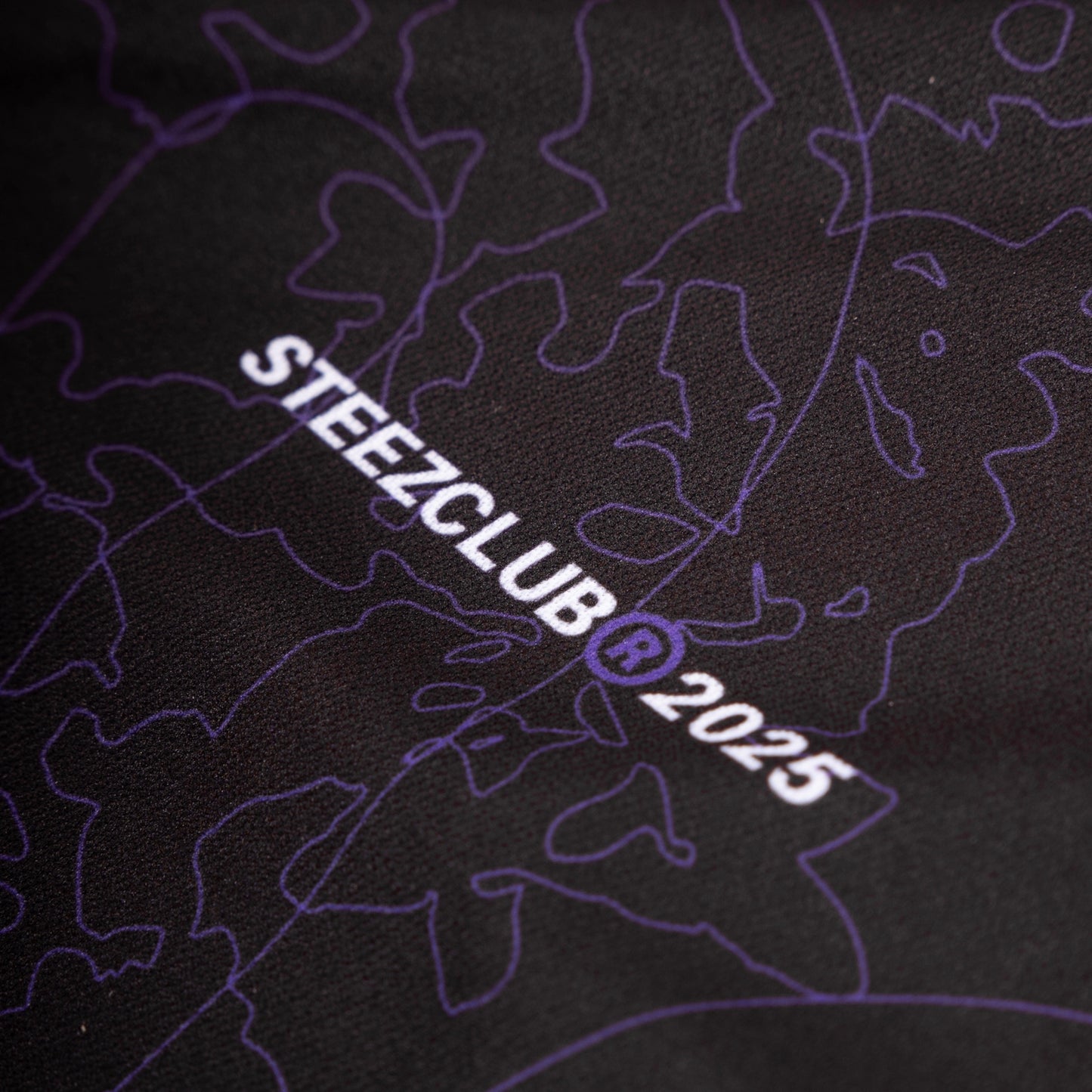 Gros plan sur un détail du maillot noir avec un imprimé graphique en ton sur ton de lignes violettes. L’inscription “STEEZCLUB ® 2025” est imprimée en blanc, avec le symbole ® en violet. L’impression semble de haute qualité, intégrée harmonieusement au design global du tissu.