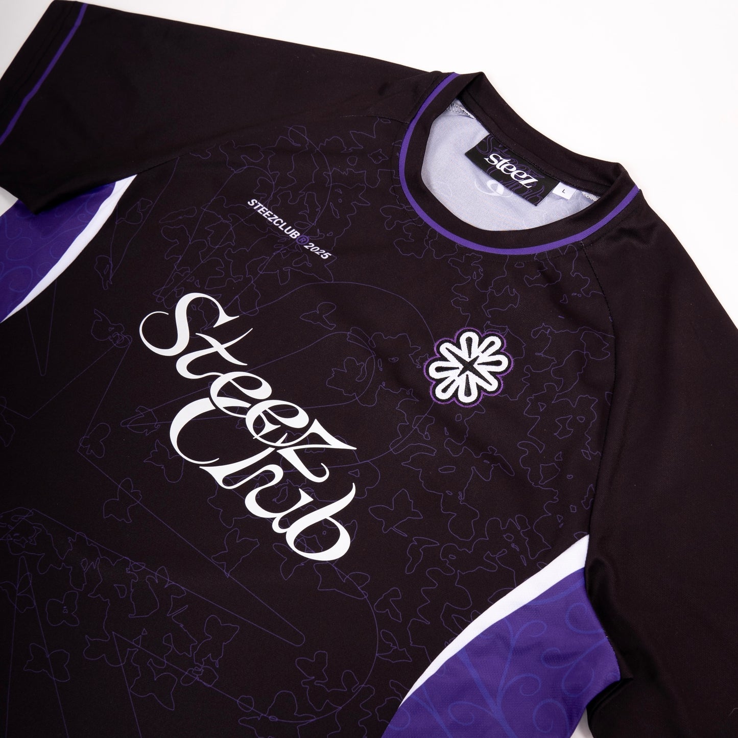 Un gros plan sur un maillot noir de la marque “Steez Club”, mettant en valeur les détails du design. Le logo “Steez Club” est imprimé en blanc sur le devant, accompagné d’un motif subtil de feuilles et de formes géométriques en ton sur ton. Un écusson brodé représentant une fleur stylisée est cousu sur la poitrine, et l’encolure est soulignée par un liseré violet. Les manches et les côtés du maillot comportent des inserts violets et blancs, ajoutant du contraste à la pièce.