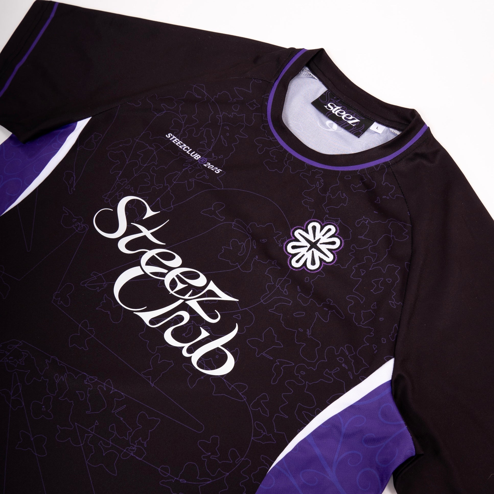 Un gros plan sur un maillot noir de la marque “Steez Club”, mettant en valeur les détails du design. Le logo “Steez Club” est imprimé en blanc sur le devant, accompagné d’un motif subtil de feuilles et de formes géométriques en ton sur ton. Un écusson brodé représentant une fleur stylisée est cousu sur la poitrine, et l’encolure est soulignée par un liseré violet. Les manches et les côtés du maillot comportent des inserts violets et blancs, ajoutant du contraste à la pièce.