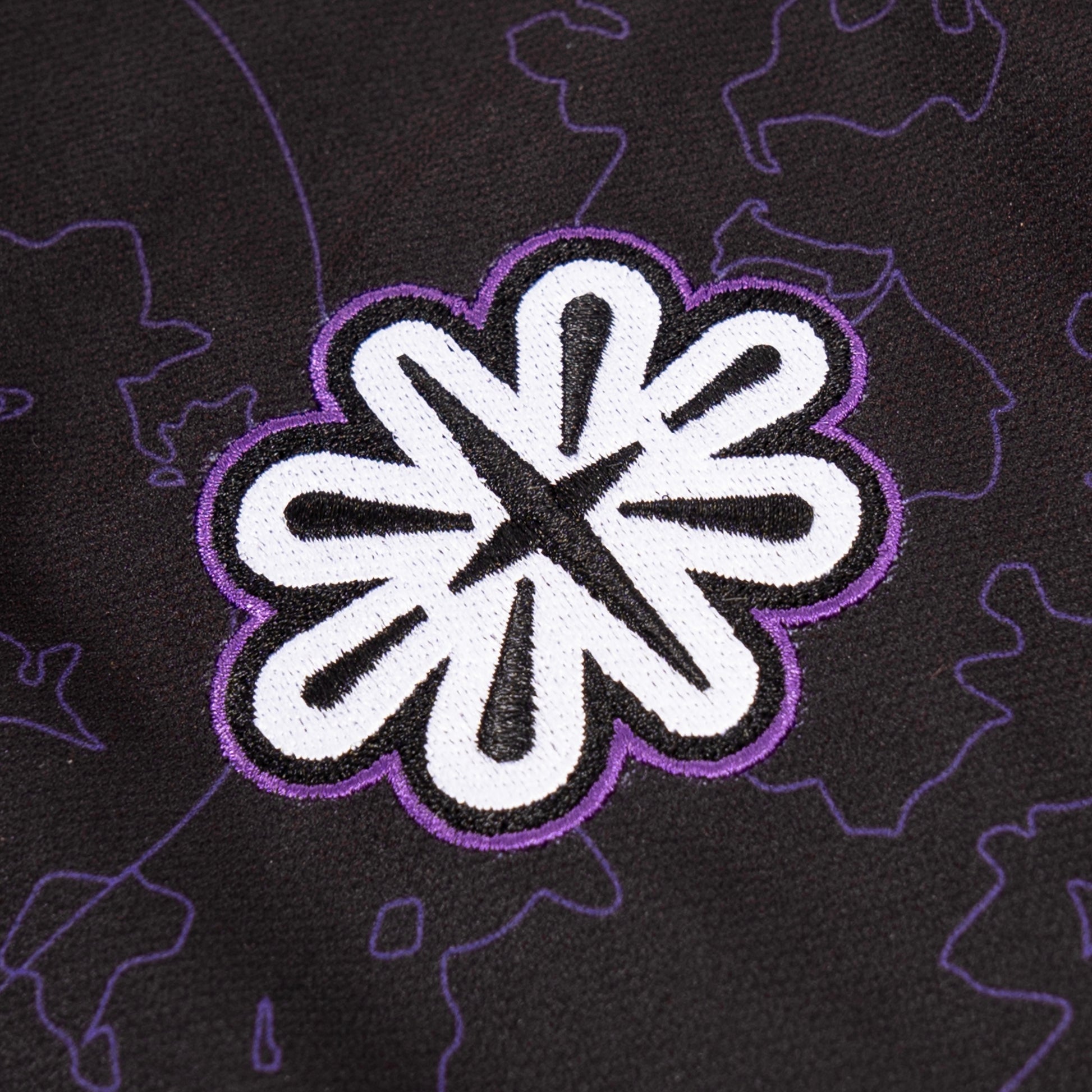 Gros plan sur un écusson brodé en forme de fleur stylisée à huit pétales, avec une bordure violette. L’intérieur de l’écusson est blanc, avec des motifs noirs qui donnent une impression de mouvement. L’écusson est cousu sur un tissu noir orné d’un motif subtil de contours violets représentant des papillons.