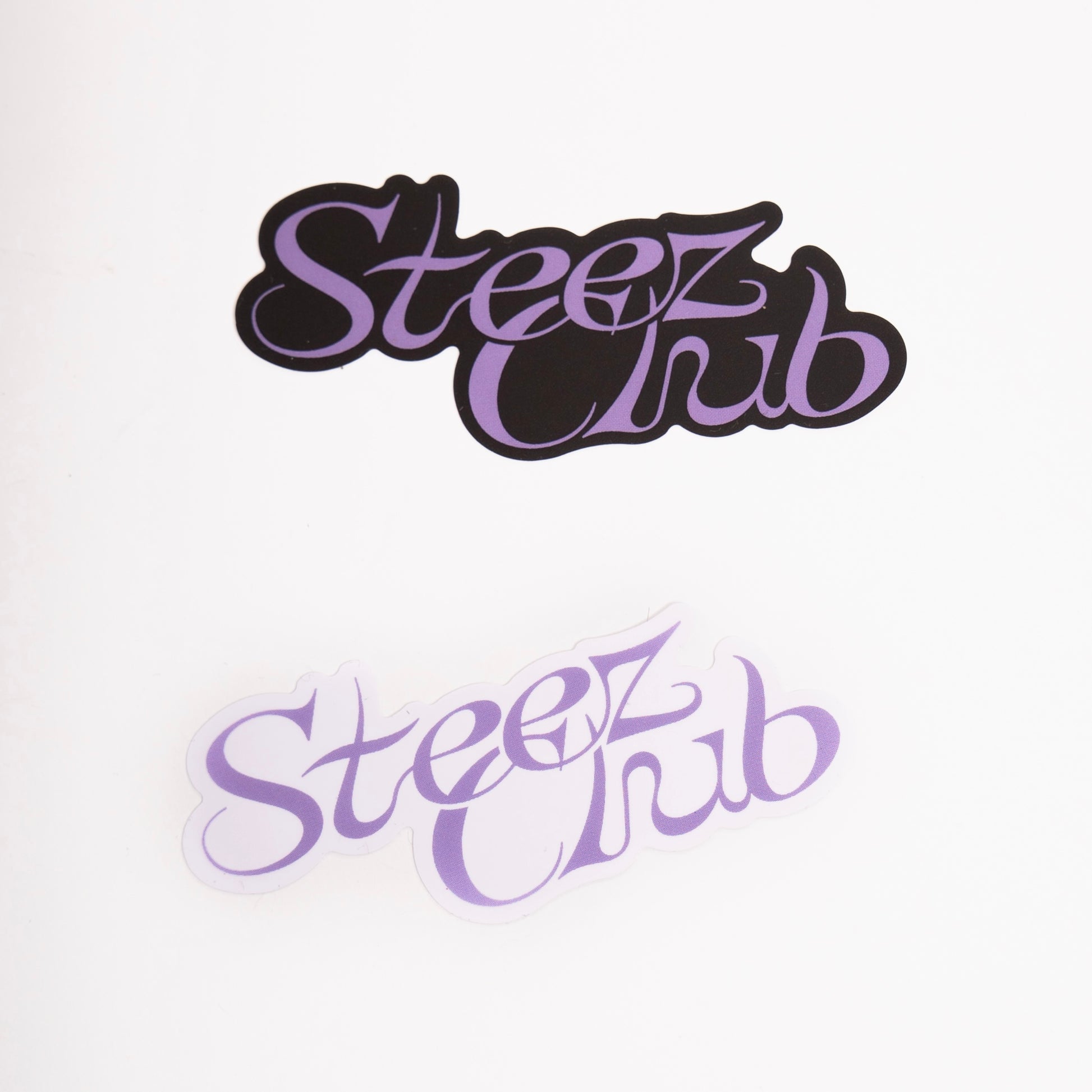 L’image montre deux stickers avec l’inscription “Steez Club” dans une typographie stylisée. L’un a un fond noir avec une écriture violette, tandis que l’autre a un fond transparent avec une écriture violette plus claire. Ils sont posés sur une surface blanche, mettant en valeur leur design.