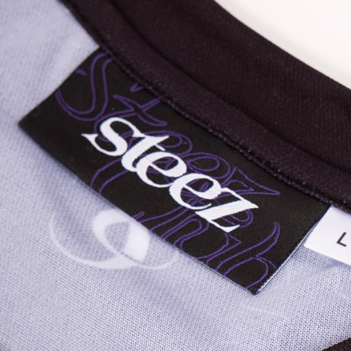 Gros plan sur l’étiquette d’un maillot de la marque “Steez Club”. L’étiquette noire arbore le logo “steez” en blanc avec un fond violet où l’on distingue le texte “Steez Club” en filigrane. Le col noir du maillot est visible, ainsi qu’une partie du tissu intérieur blanc et violet. Une étiquette blanche indiquant la taille “L” est également cousue à côté du label principal.