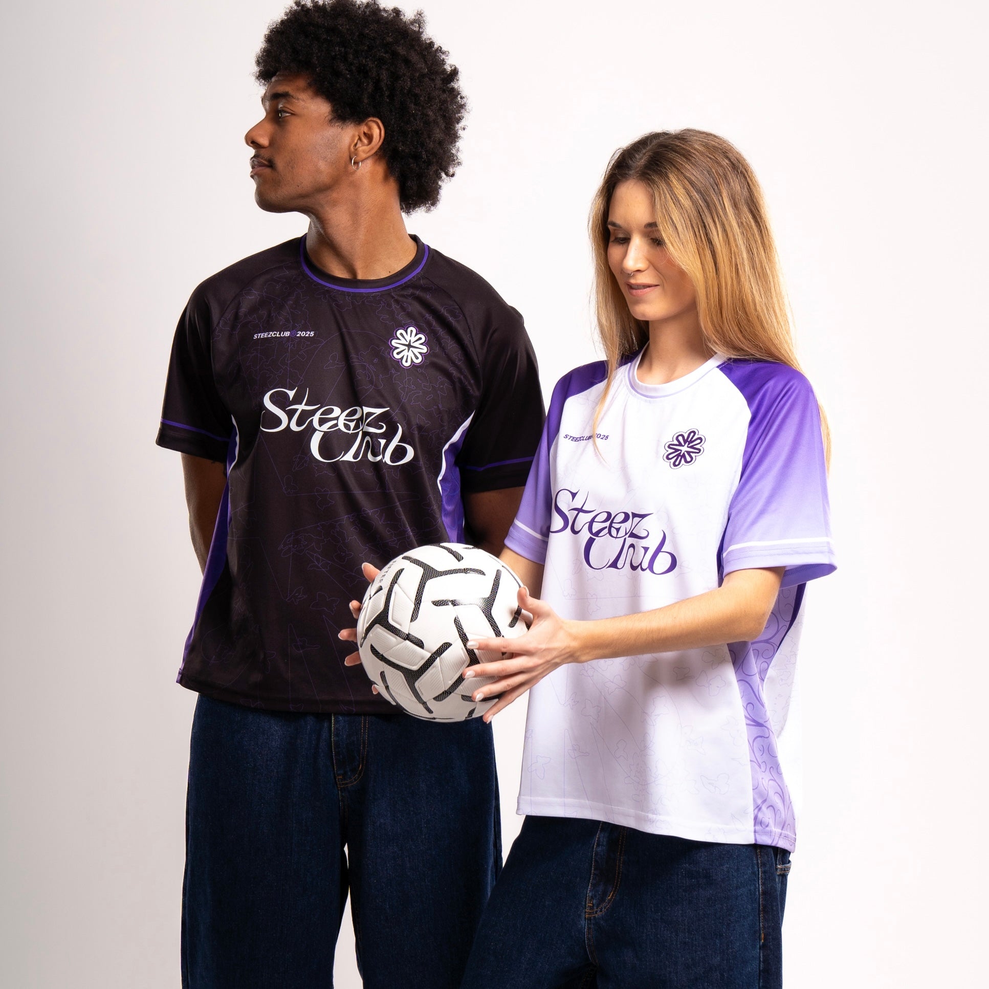 Deux mannequins posent en portant des maillots de football de la marque "Steez Club". L'homme, à gauche, porte un maillot noir avec des détails violets et blancs, tandis que la femme, à droite, porte un maillot blanc avec des manches violettes. Ils sont habillés en jeans et adoptent une posture confiante. La femme tient un ballon de football blanc et noir dans ses mains. L'arrière-plan est blanc et minimaliste