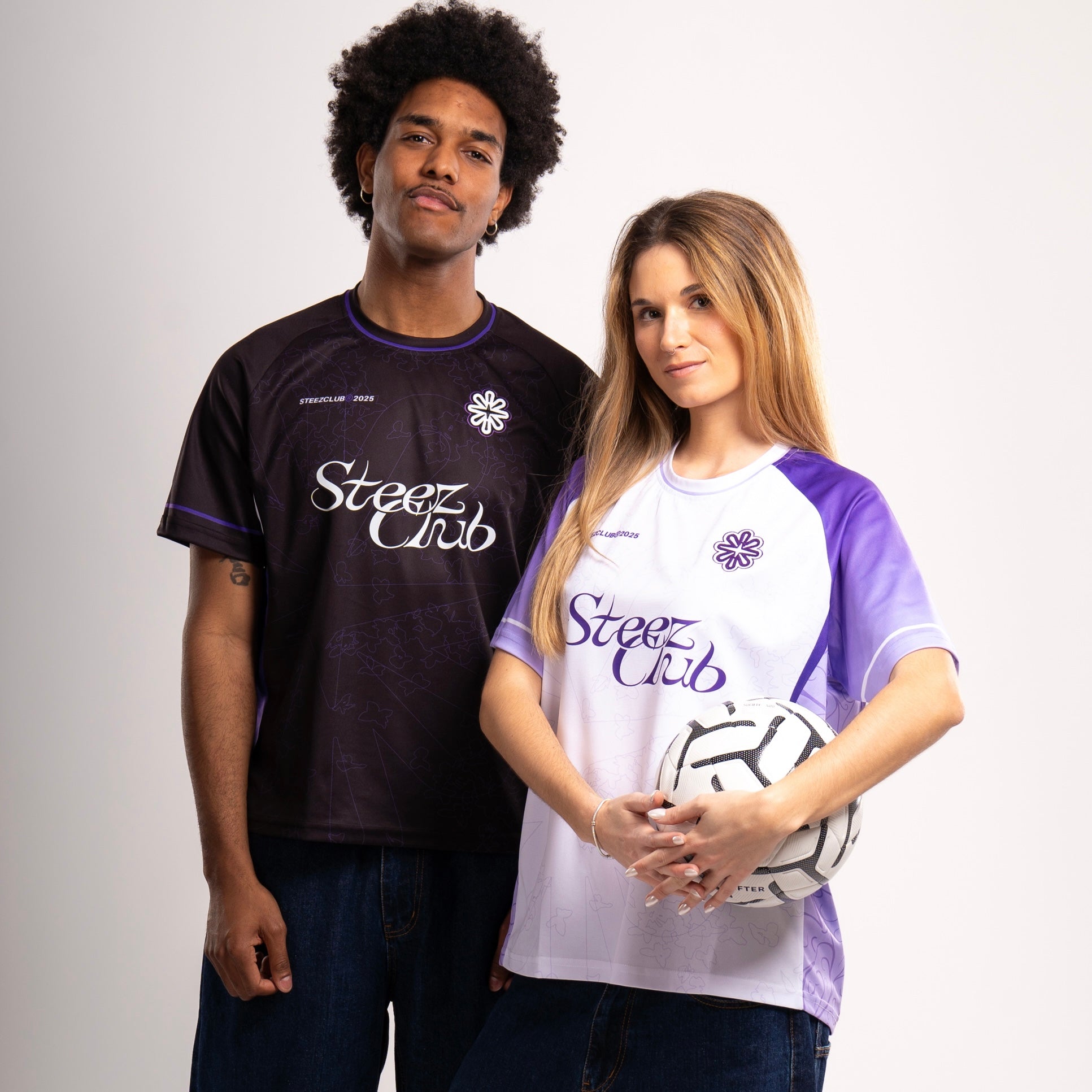 Deux modèles posent ensemble en portant les maillots de la collection “Steez Club”. L’homme porte la version noire avec des détails violets, tandis que la femme porte la version blanche avec des manches violettes. Les deux maillots arborent le logo “Steez Club” sur la poitrine ainsi qu’un motif subtil en ton sur ton. La femme tient un ballon de football entre ses mains, renforçant l’esthétique sportive de la tenue.