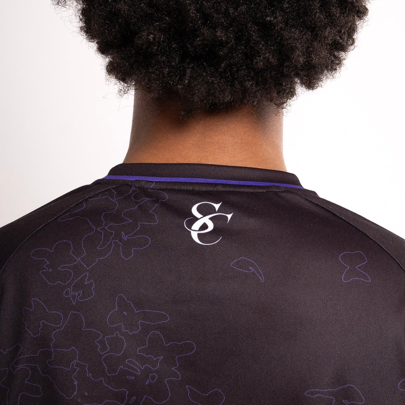 Vue arrière d’une personne portant un maillot noir de la marque “Steez Club”. Le col est souligné d’un liseré violet. Au centre du haut du dos, un logo “SC” blanc est imprimé. Le tissu du maillot est orné d’un motif subtil de papillons dessinés en contour violet. Les épaules et le haut du dos de la personne sont visibles, ainsi que la texture de ses cheveux bouclés.