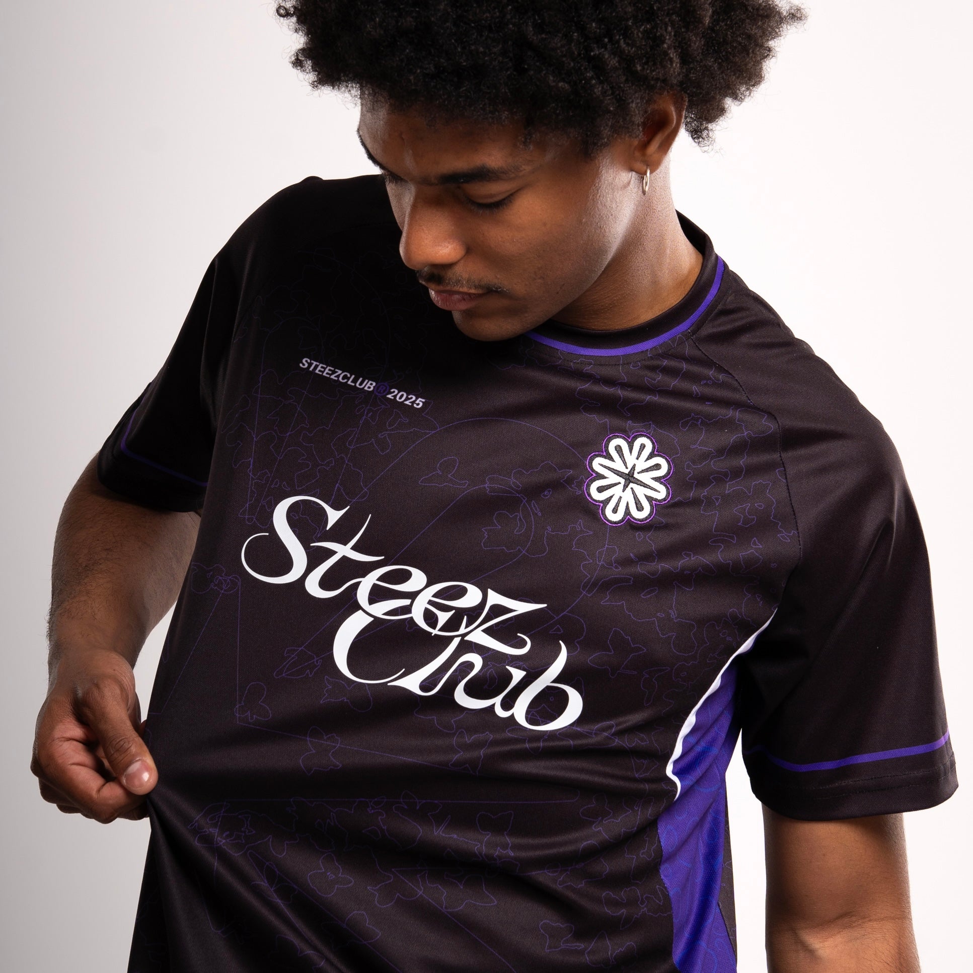 Un jeune homme aux cheveux bouclés porte un maillot noir de la marque “Steez Club”. Il regarde vers le bas en tirant légèrement sur le tissu, mettant en avant les détails du design. Le logo “Steez Club” est imprimé en blanc sur la poitrine, accompagné d’un motif subtil de feuilles et de formes géométriques ton sur ton. Un écusson brodé représentant une fleur stylisée est cousu sur le côté gauche du torse. L’encolure et les manches sont ornées de liserés violets, contrastant avec le noir du maillot.