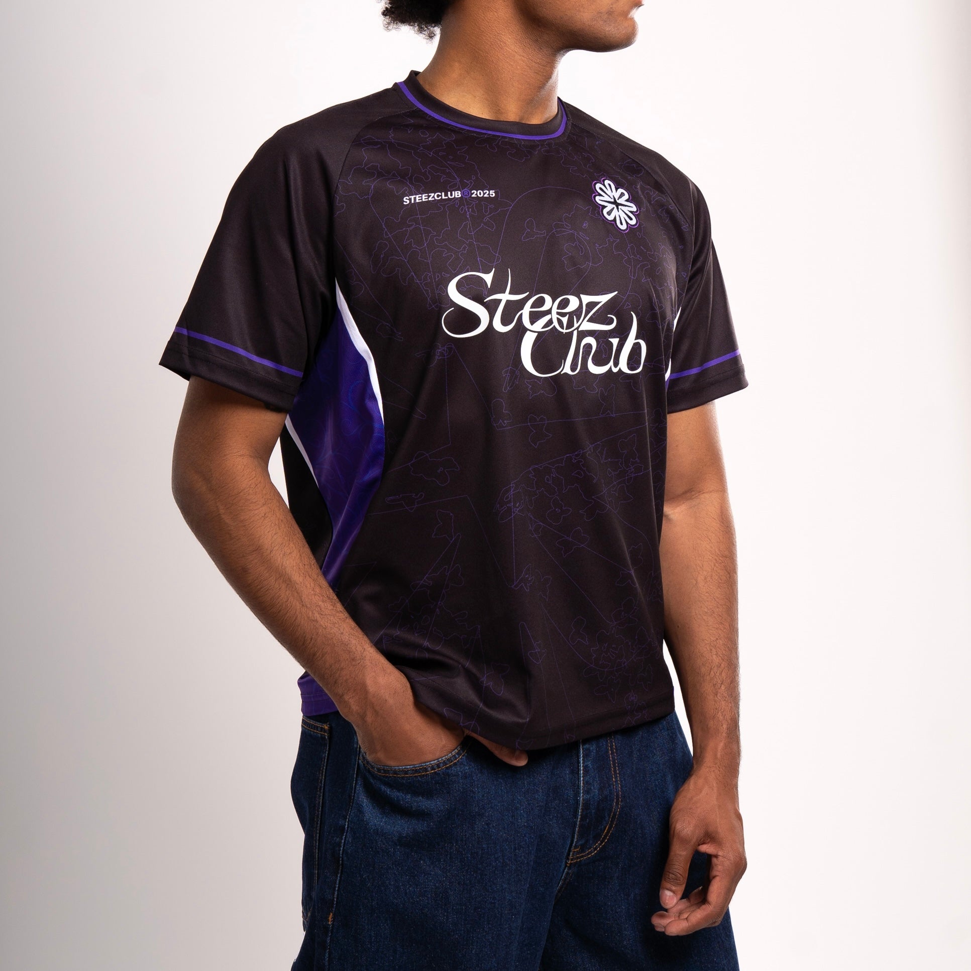 Un homme porte un maillot noir à manches courtes avec des accents violets et blancs. Le maillot présente un design subtil en ton sur ton avec des motifs de papillons et arbore l’inscription “Steez Club” en blanc sur la poitrine. Un petit écusson brodé en forme de fleur stylisée est cousu sur le haut du torse. L’inscription “STEEZCLUB ® 2025” est visible sur le côté gauche du maillot. L’homme associe le maillot à un jean ample et adopte une posture décontractée, une main dans la poche.